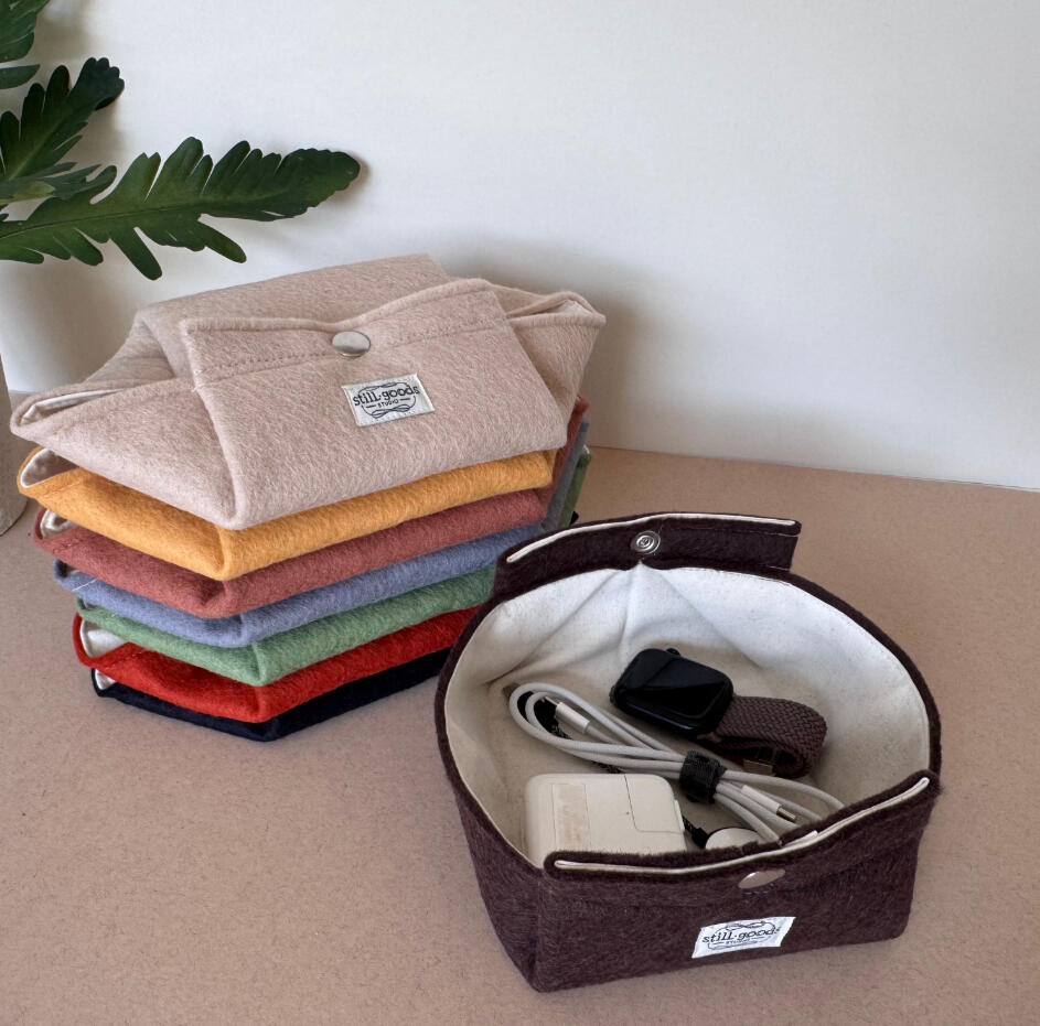 Stack of folded wool felt pouches in an array of colors, beside an open round felt caddy holding a charging cable, styled with a green plant leaf.