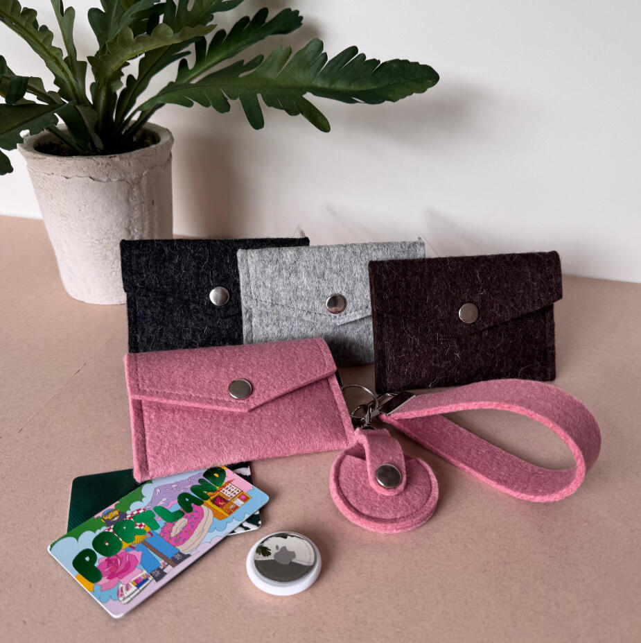 EDC Bundle Snap-close wool felt card wallets in charcoal, grey, and pink, alongside a wool AirTag holder and wrist strap, with Portland postcards on the table.