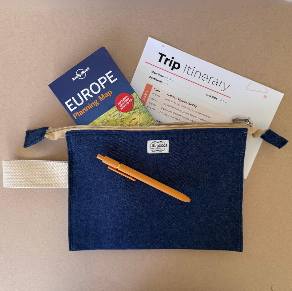 Navy cotton zip pouch with the StillGoods Studio label, holding a travel itinerary and Europe guidebook, with a pen tucked into its canvas strap.