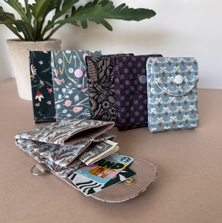 Snap-close fabric wallets in floral and botanical prints, opened to show cards tucked inside, styled with a small potted plant.